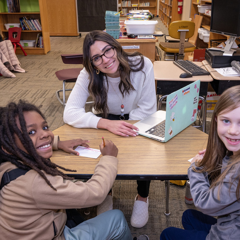 student teacher with laptop sitting at table with two smiling elementary students
