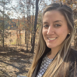 Woman with long brown hair standing outside with trees in the background during fall.