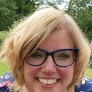 Woman with chin-length blond hair and glasses smiling and posing outside with trees and green grass behind her