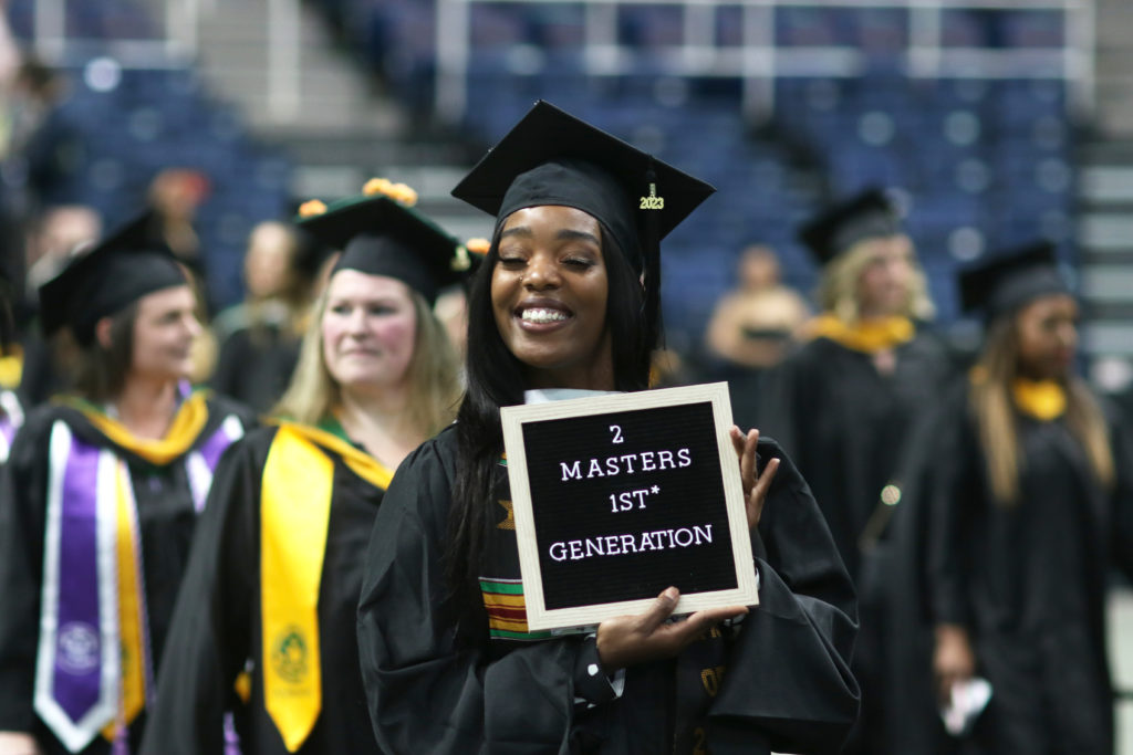 2023 graduate in cap and gown holding sign that says, "2 Master's 1st Generation"