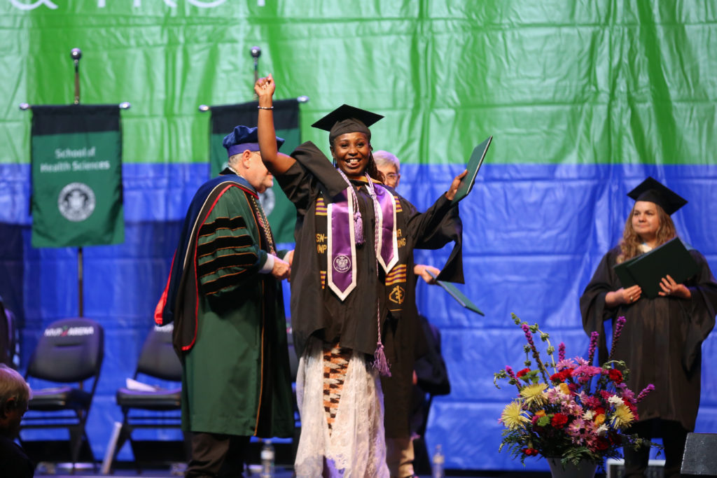 A graduate on commencement stage raises arms with diploma in hand