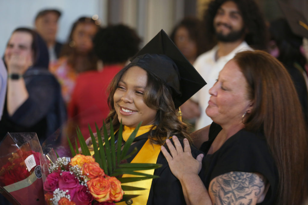 Woman in a graduation cap and gown holds flowers and receives hug from second woman