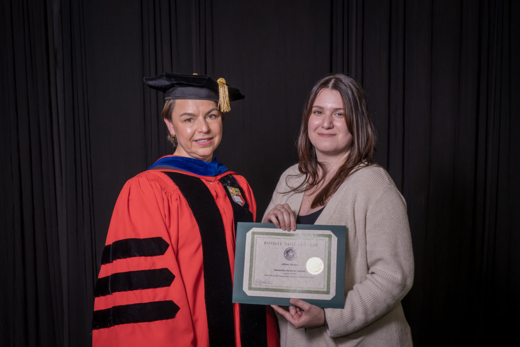 woman in red graduation gown and black hat standing with a female college student with long brown hair and a tan cardigan over a black top holding certificate