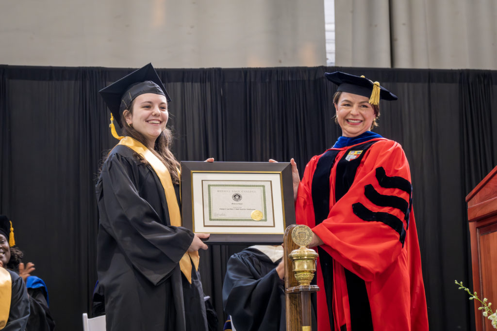 woman in red graduation gown and black graduation cap holding one side of a large framed certificate while female college student in black graduation cap and gown with shoulder length brown hair holds other side of plaque
