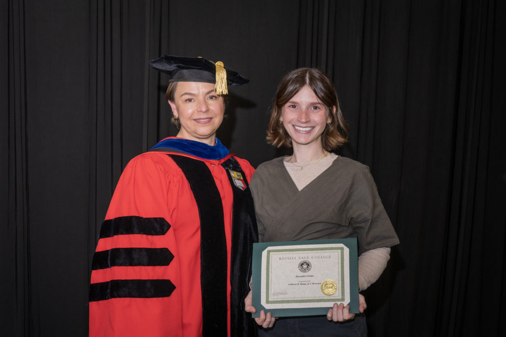 woman in red graduation gown and black hat standing with a female college student with chin-length brown hair holding certificate