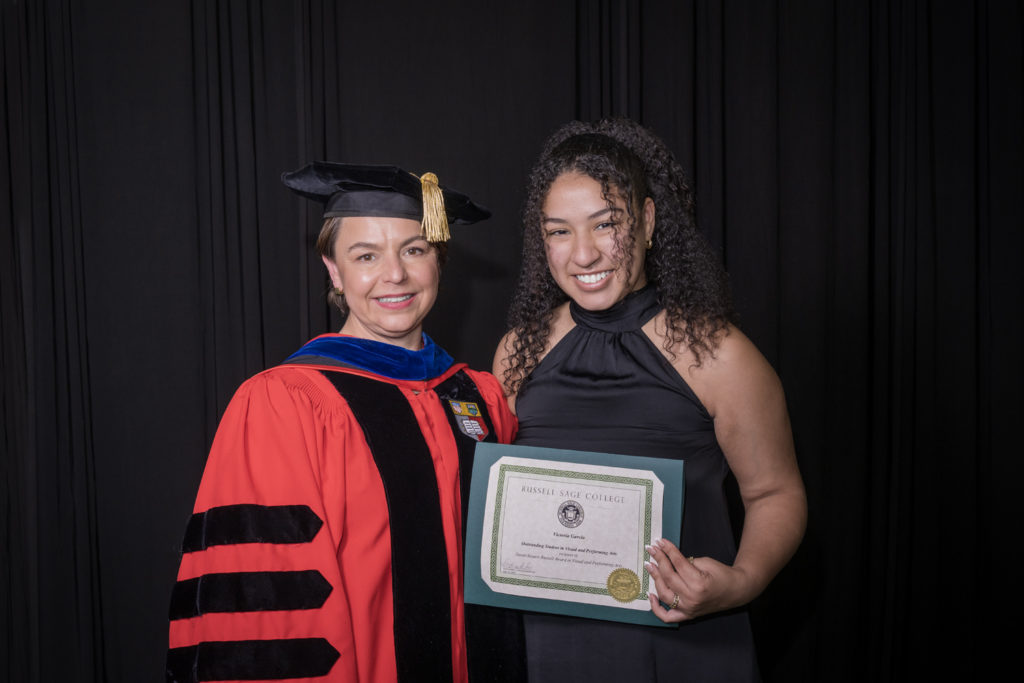 woman in red graduation gown and black hat standing with a female college student with curly brown hair holding certificate