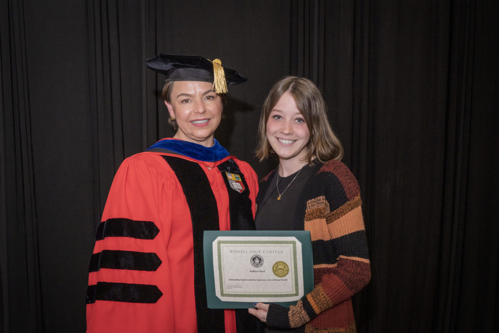 woman in red graduation gown and black hat standing with a female college student with long brown hair and a striped sweater over a black dress holding certificate