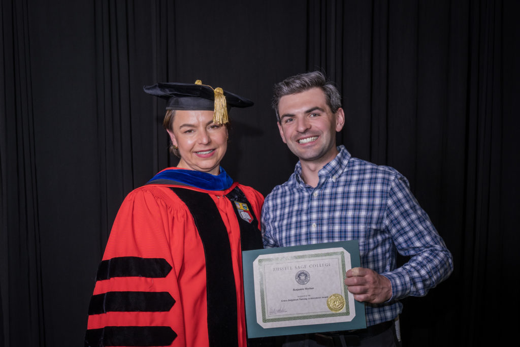 woman in red graduation gown and black hat standing with a male college student in blue checkered dress shirt holding certificate