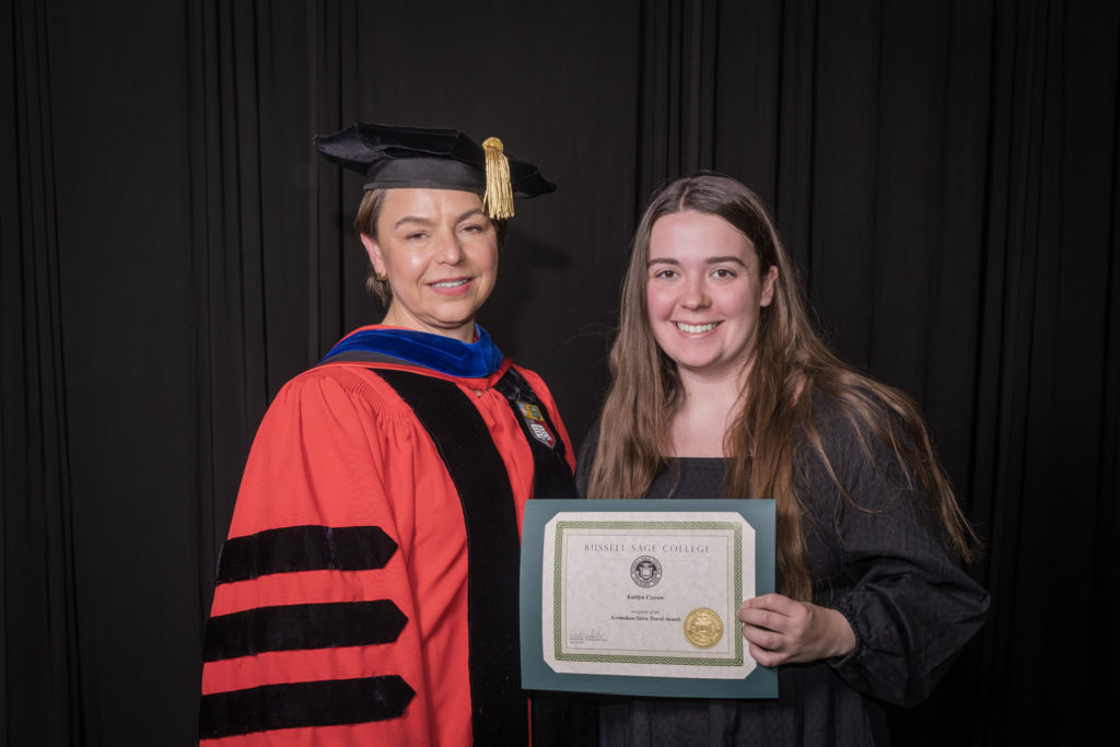 woman in red graduation gown and black hat standing with a female college student with long brown hair and a black dress holding certificate