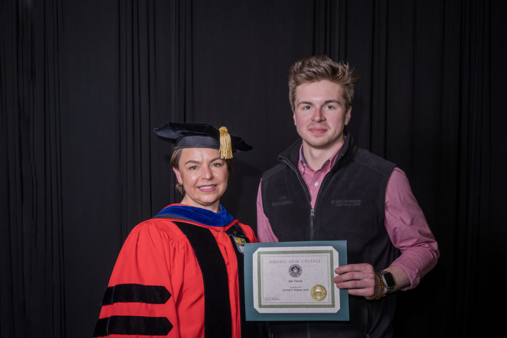 woman in red graduation gown and black hat standing with a male college student wearing a red dress shirt and a gray vest holding certificate