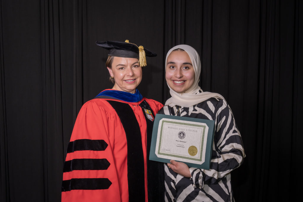 woman in red graduation gown and black hat standing with a female college student wearing a tan hijab holding certificate