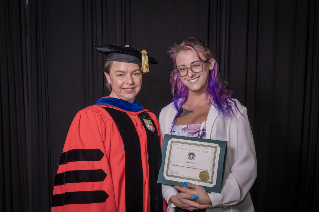 woman in red graduation gown and black hat standing with a female college student with purple and pink shoulder length hair  and a white jacket over a floral dress holding certificate