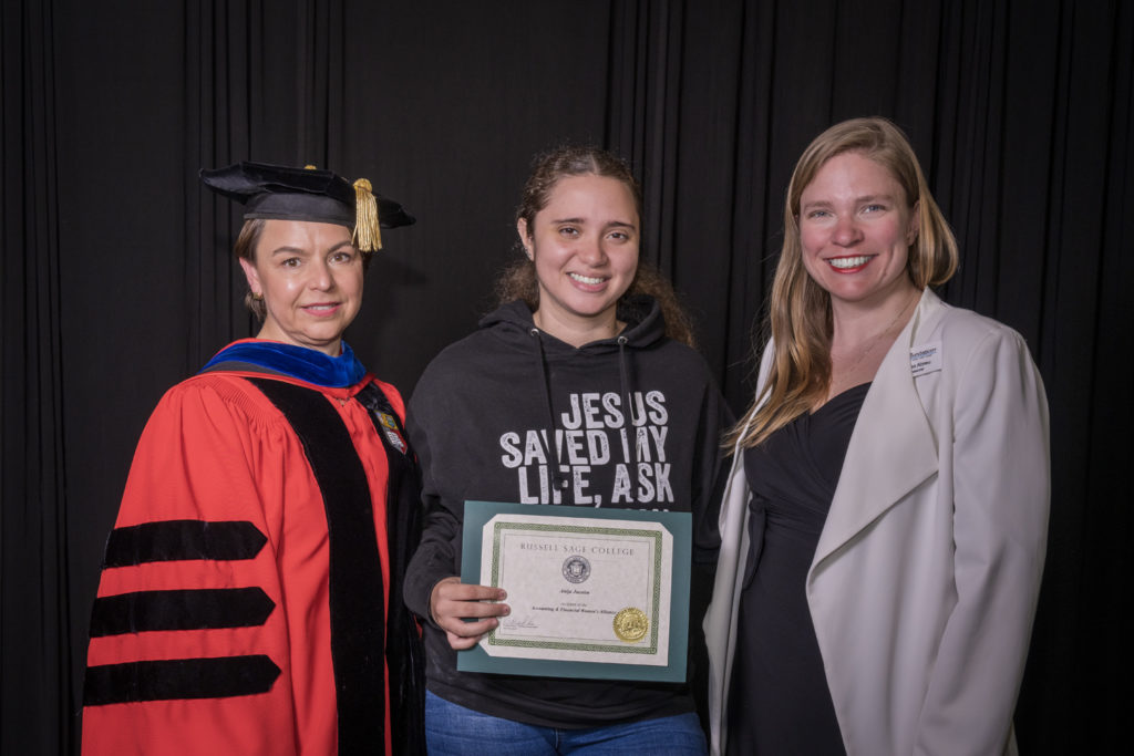 woman in red graduation gown and black hat standing with a female college student wearing a sweatshirt saying Jesus saved my life holding certificate and standing next to a woman in a white coat with long blond hair