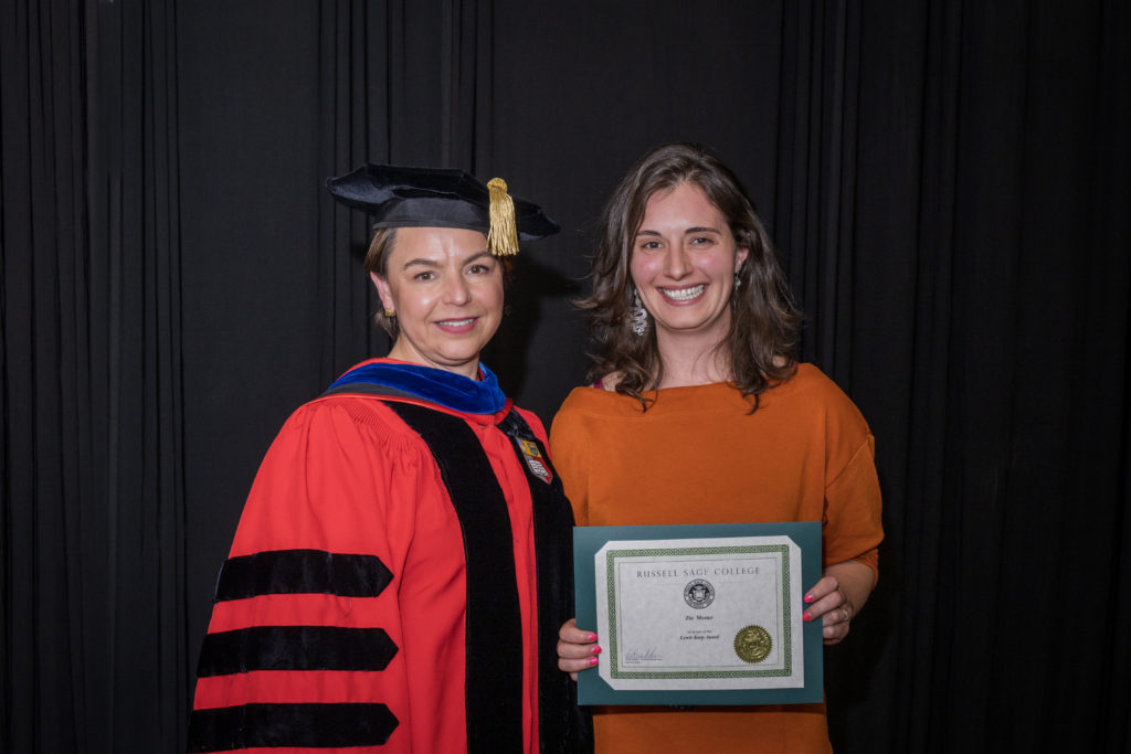 woman in red graduation gown and black hat standing with a female college student with shoulder-length brown hair and an orange shirt holding certificate