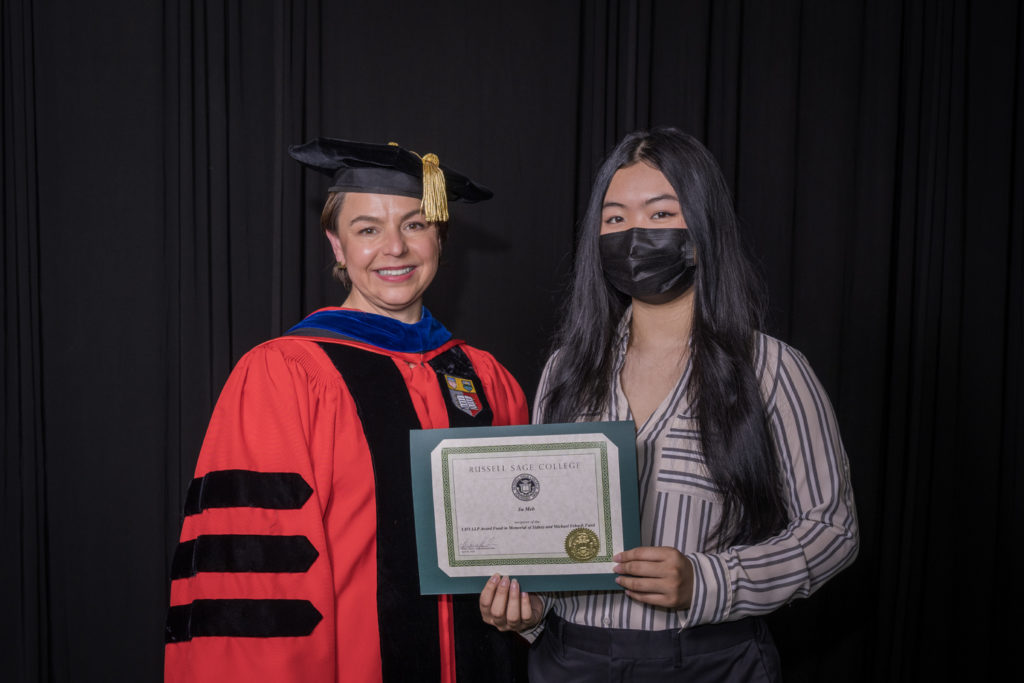woman in red graduation gown and black hat standing with a female college student with long black hair and wearing a black surgical mask holding certificate