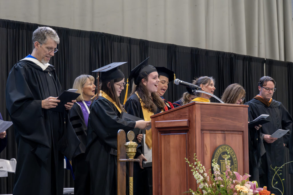 College students and faculty dressed in commencement gowns singing behind a podium.