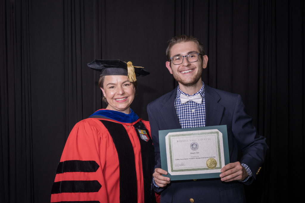 woman in red graduation gown and black hat standing with a male college student in a blue checkered shirt, blue jacket and white bow tie holding certificate