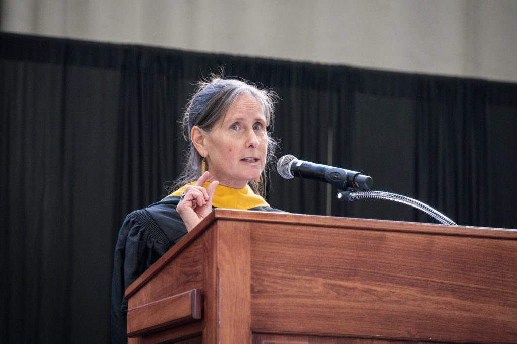 Middle-aged woman with hair pulled back in graduation gown speaking behind podium