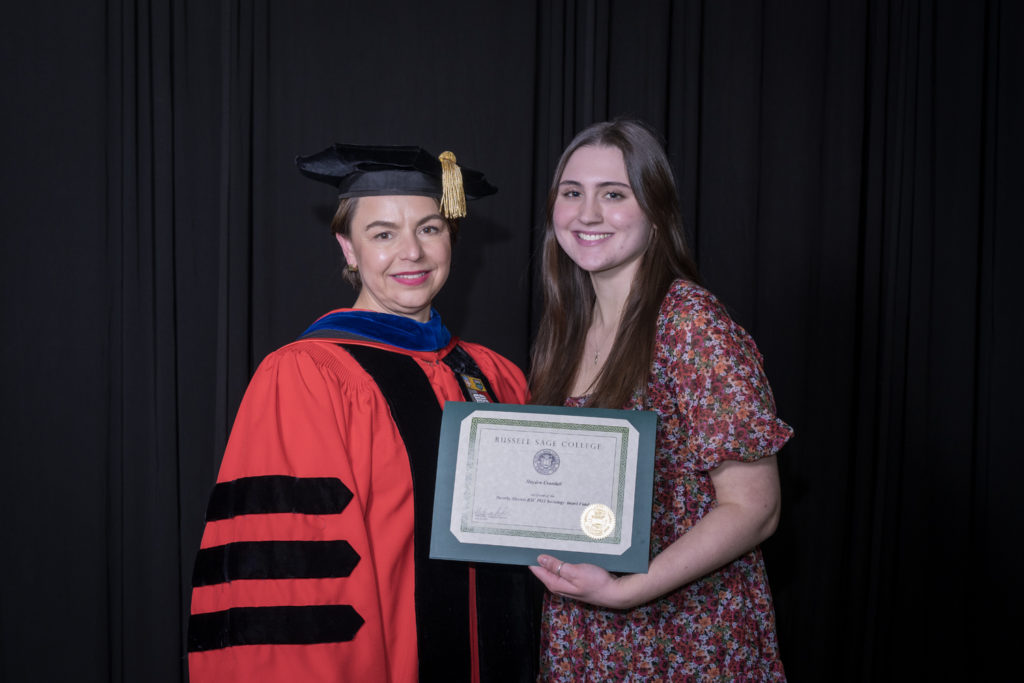 woman in red graduation gown and black hat standing with a female college student with long brown hair holding certificate