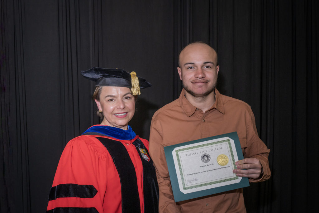 woman in red graduation gown and black hat standing with a male college student with a brown dress shirt and slight beard holding certificate