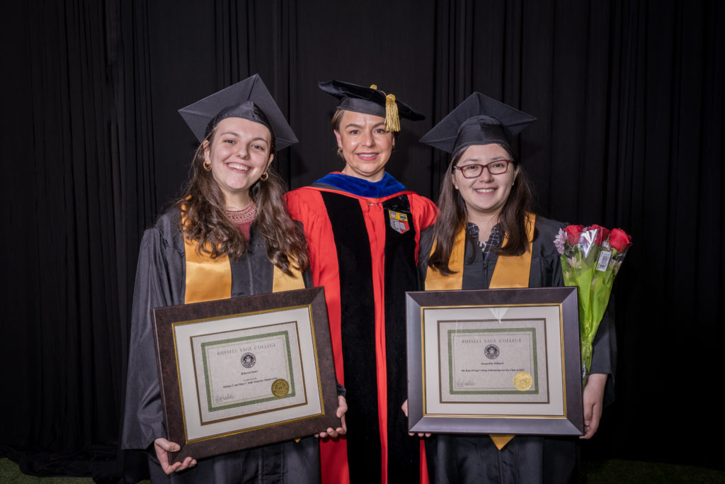 woman in red graduation gown and black hat standing with two female college students in black commencement caps and gowns holding certificates