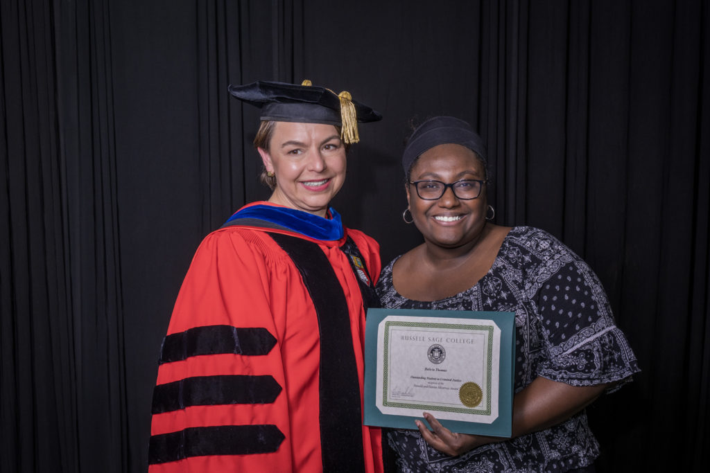 woman in red graduation gown and black hat standing with a female college student wearing a headband and glasses holding certificate