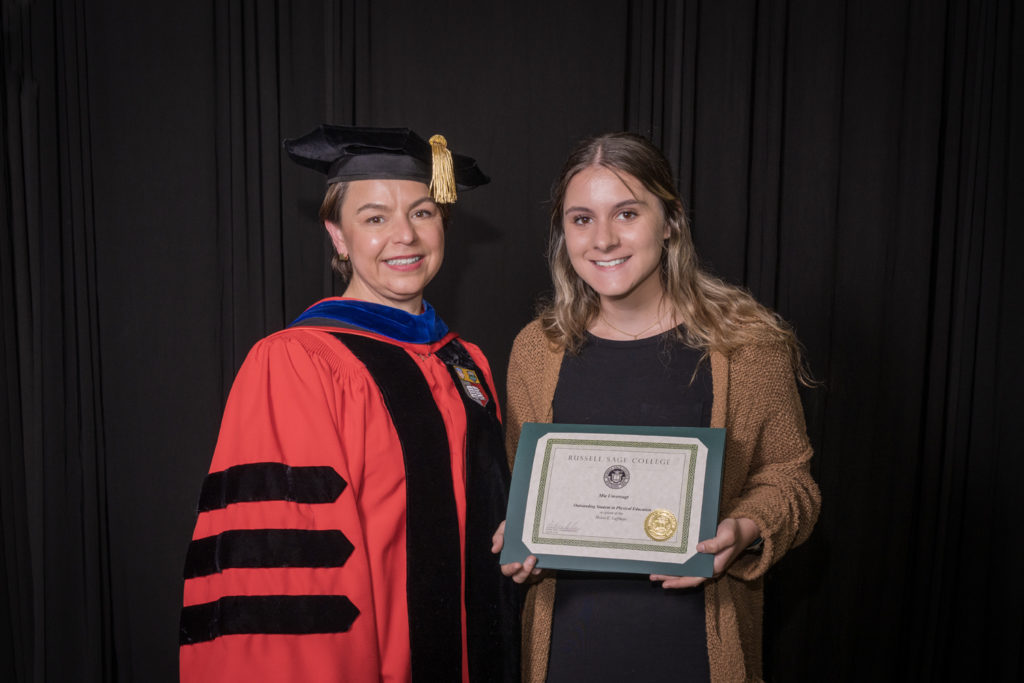 woman in red graduation gown and black hat standing with a female college student with long blond hair and a black dress with a camel colored cardigan holding certificate