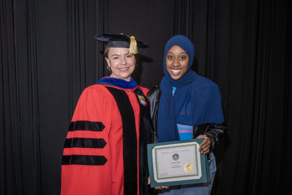 woman in red graduation gown and black hat standing with a female college student in blue hijab holding certificate