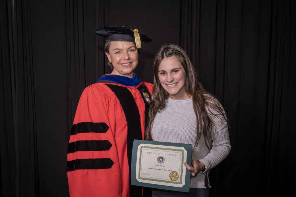 woman in red graduation gown and black hat standing with a female college student with long blond hair  and a tan long-sleeve shirt holding certificate