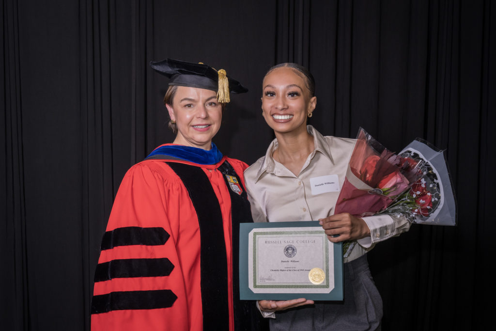 woman in red graduation gown and black hat standing with a female college student with hair pulled back in ponytail holding flowers and a certificate