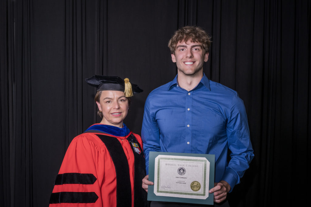 woman in red graduation gown and black hat standing with a male college student in blue dress shirt holding certificate