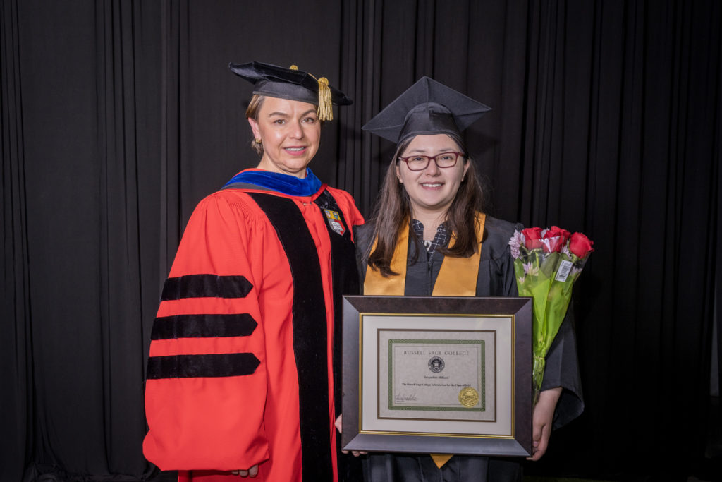 woman in red graduation gown and black hat standing with a female college student also in commencement gear holding flowers and a certificate