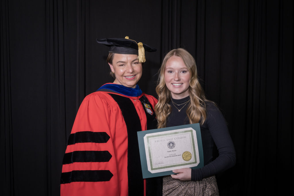 woman in red graduation gown and black hat standing with a female college student with long blond hair holding certificate