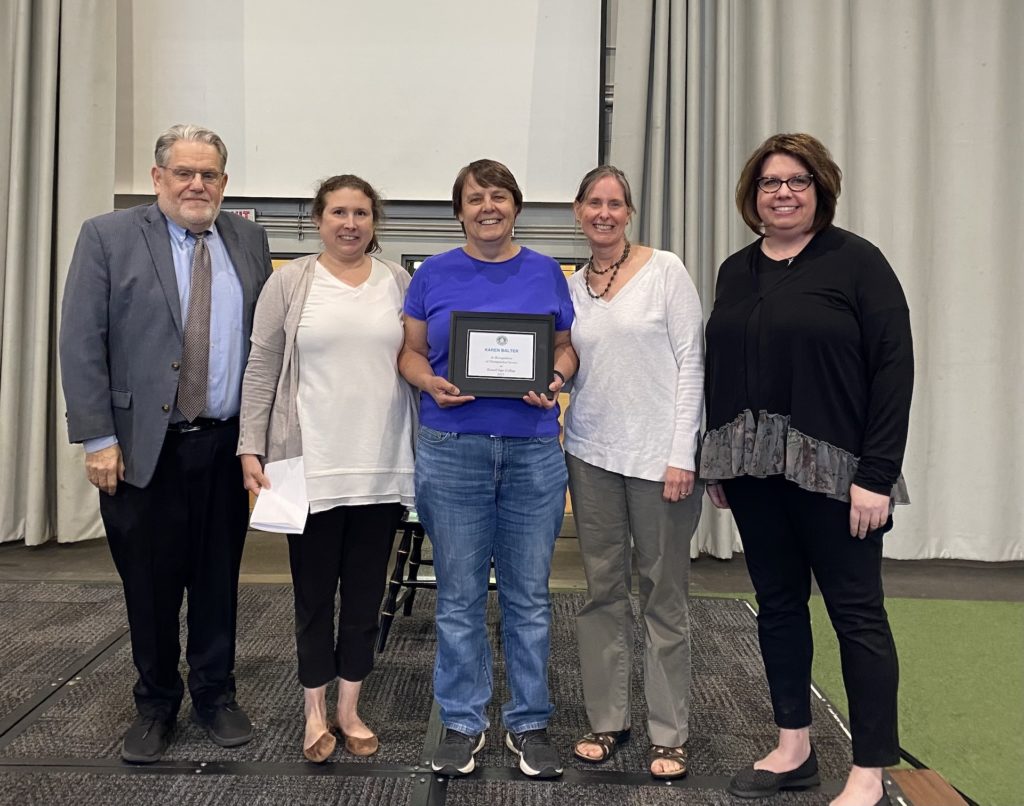 Karen Balter, associate professor and chair of the biology and health sciences program, standing at center with a certificate surrounded by President Christopher Ames and three female faculty colleagues.