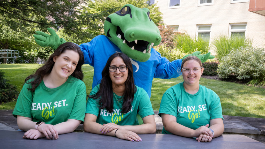 outdoor shot of three female students seated at picnic table with alligator mascot behind them