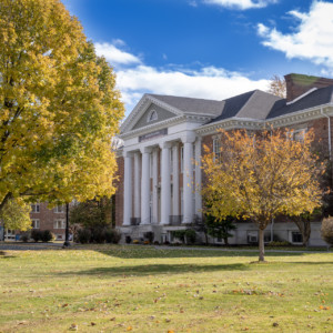 large brick building with white pillars with large swath of green grass in front of it, a two golden yellow trees to the left and in front of the building and blue skies above.