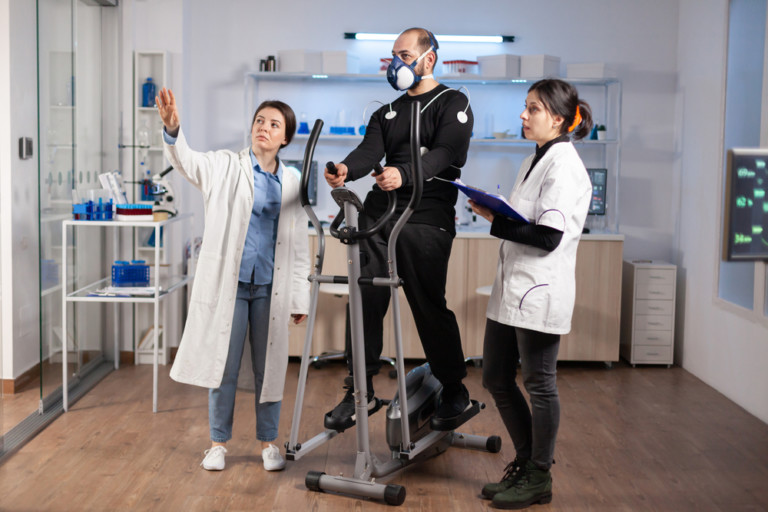 Two doctors standing in a sports performance lab working with a patient who is on a stationary bike with a breathing mask on.