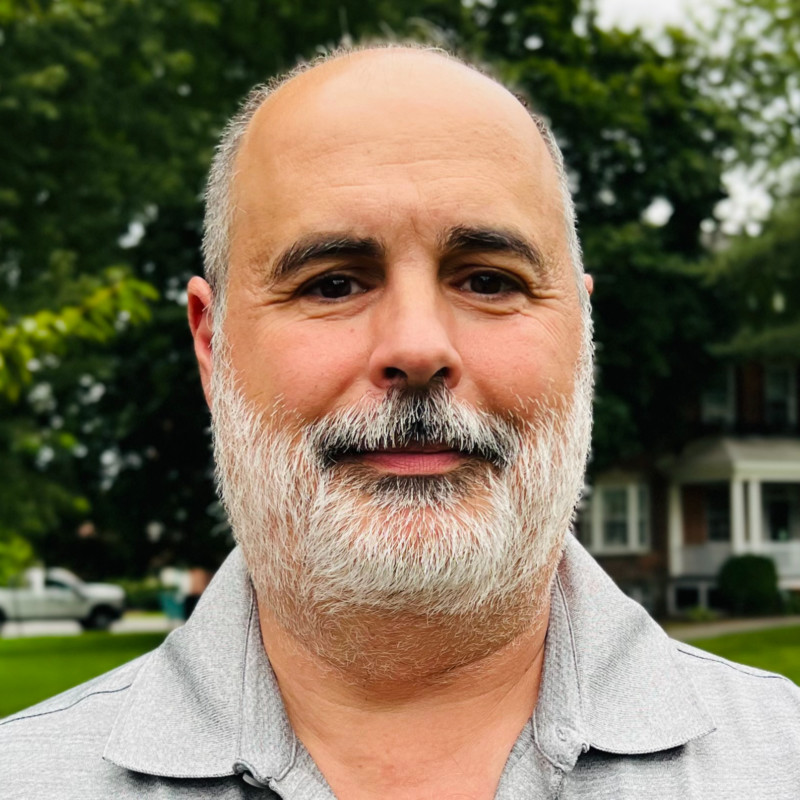 man with a white beard and a gray polo shirt standing outdoors.