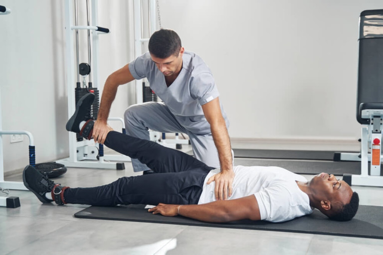 A male physical therapist in scrubs helps a male patient in a white shirt and black pants perform a lower body stretch