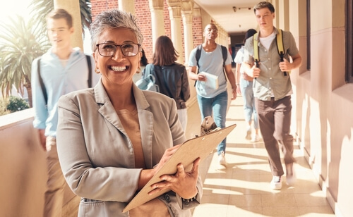 A female school administrator holds a clipboard as students walk down a hallway behind her