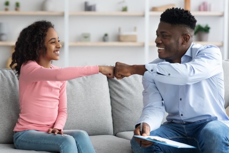 A man and girl sit on a gray couch and smile at each other while they fist bump; the man is holding a clipboard 