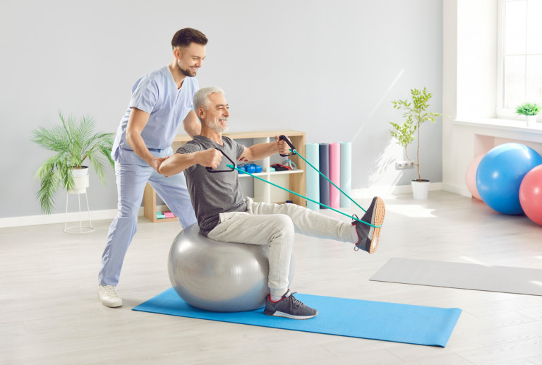 A male physical therapist in scrubs helps an older man stretch while sitting on a blue yoga ball with a resistance band around his foot