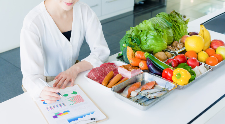 A dietitian holds a pen over a printout of charts on a white table; next to the charts are trays of fresh food, including raw meat, fish, lettuce, vegetables, and fruits