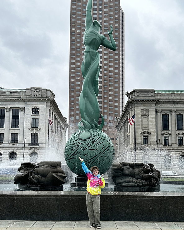 A tall green statue with an upward-reaching figure atop a patterned sphere among fountains, with buildings in the background and a person in a tie-dye hoodie pointing at it.