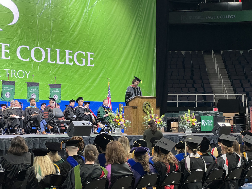 Man, dressed in cap and gown, speaking behind podium at Russell Sage College graduation ceremony
