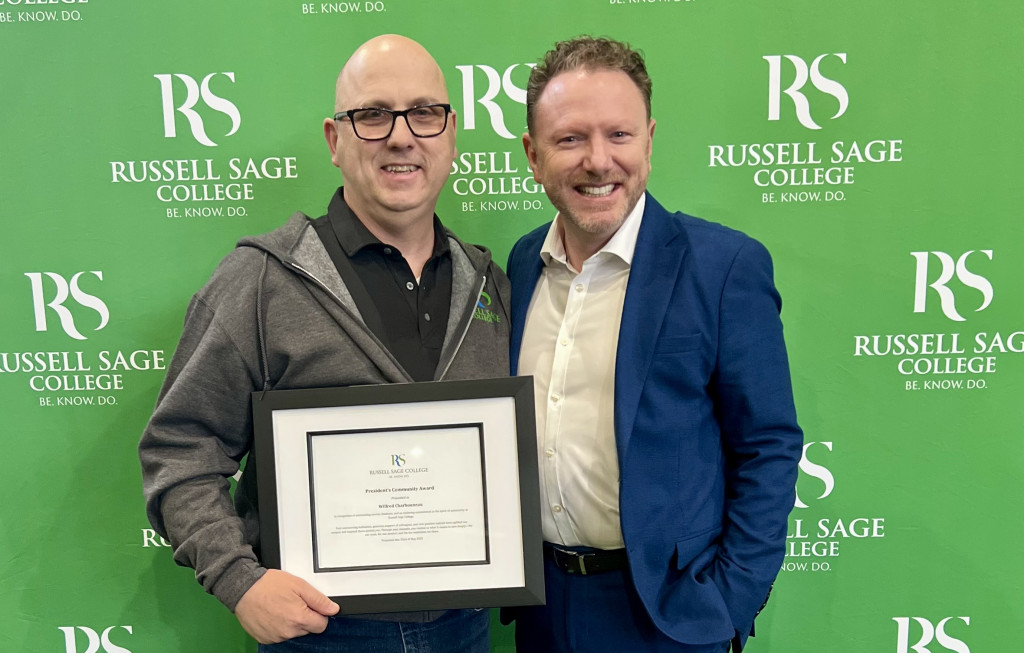 two people standing in front of green background with Russell Sage logo smiling at the camera. One person is holding a certificate.