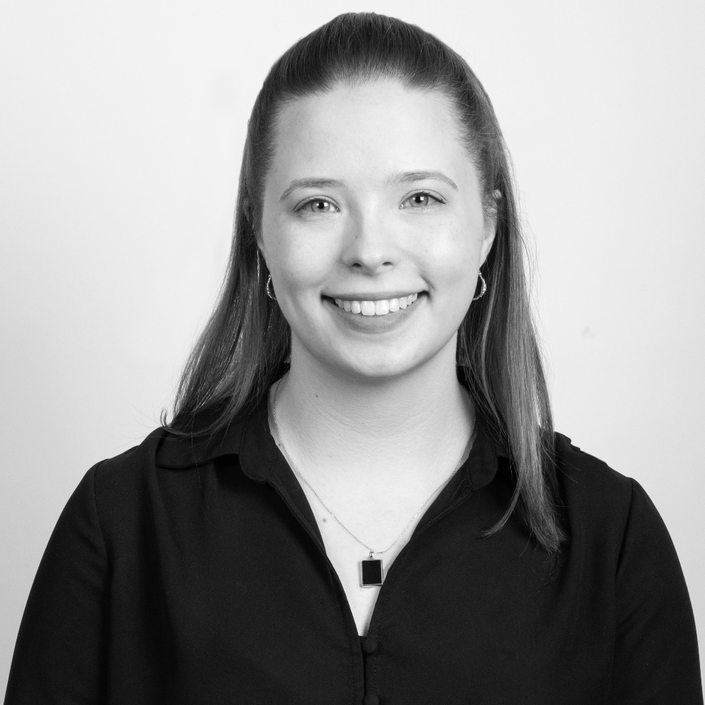 Smiling woman in a black shirt with a pendant necklace. She has long hair pulled back and hoop earrings, set against a plain white background. Black and white photo.