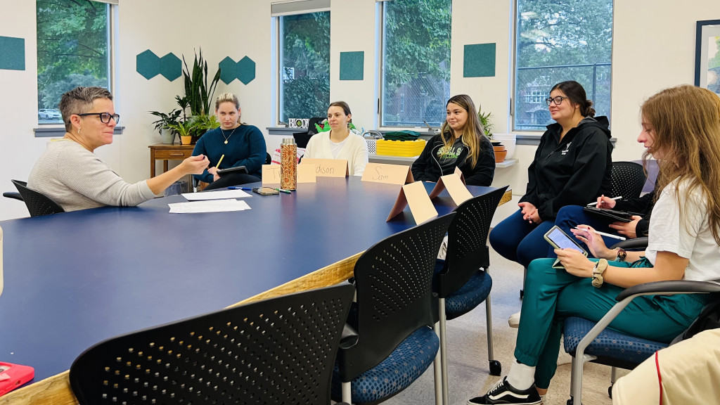 A group of people in a meeting room seated around a blue table, with one person speaking and others listening.