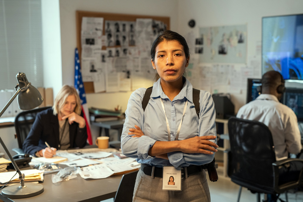 A woman in the criminal justice field stands confidently in an investigative office, with colleagues working in the background.