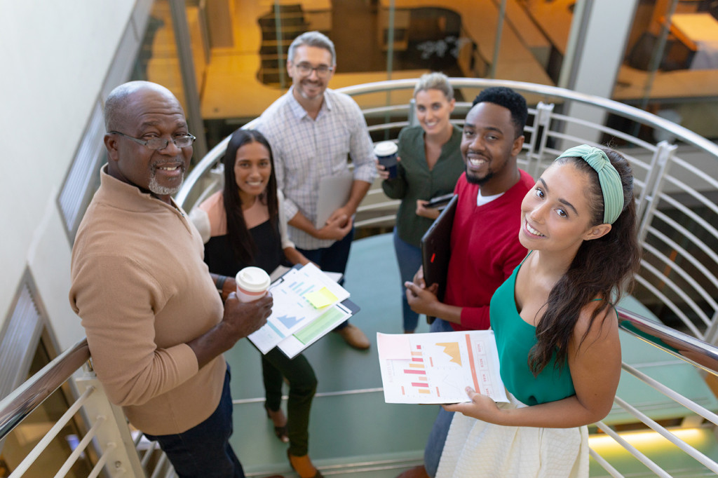 Six people on a curved staircase in a school setting, holding documents and coffee cups, looking up at the camera.